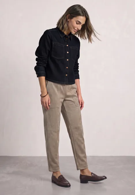 Femme porte veste en jean noire, pantalon beige et mocassins bruns, debout sur fond neutre, regard baissé.