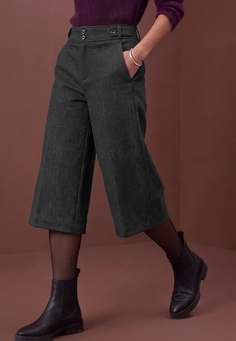 Bermuda met High Waist en Wide Leg pijpen in casual pasvorm Black Dunkle Culottes kombiniert mit schwarzen Stiefeletten und zarten Strumpfhosen, vor rotem Hintergrund.