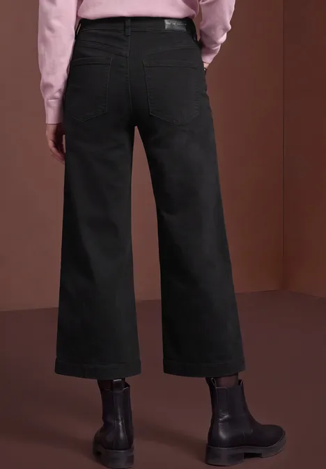 Mid Waist Jeans Culotte im Loose Fit black authentic washed Rückenansicht einer Person in schwarzen, weiten Jeans und schwarzen Stiefeln vor braunem Hintergrund.