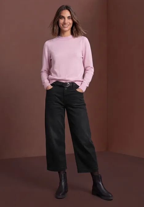 Mid Waist Jeans Culotte im Loose Fit black authentic washed Lächelnde Frau in rosa Pullover, schwarzen Culotte-Hosen und Stiefeln vor braunem Hintergrund.