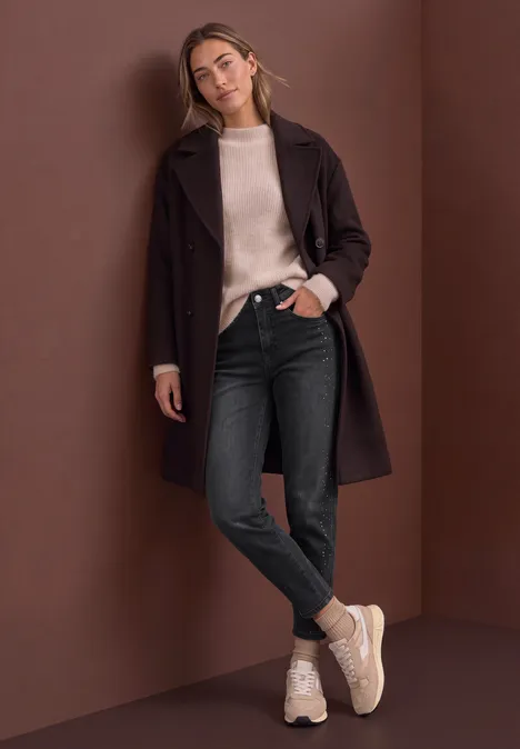 High Waist Barrel Leg Jeans im Loose Fit grey washed Frau lehnt lässig an brauner Wand, trägt braunen Mantel, beigen Pullover, schwarze Jeans und helle Sneaker.