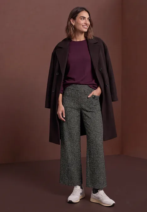 Femme en pull bordeaux, manteau sombre, pantalon large gris et baskets blanches devant un mur marron uni.