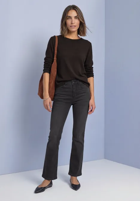 Bootcut-jeans met Mid Waist-taille in Slim Fit Black Washed Frau in schwarzem Langarmshirt, schwarzen Bootcut-Jeans, Pumps und brauner Tasche vor pastellfarbenem Hintergrund.