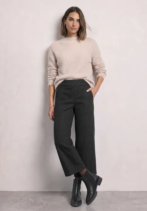 Culotte met High Waist en Loose Fit-pasvorm Black Frau in hellem Pullover, dunkelgrauen, weiten, cropped Hosen und schwarzen Boots vor hellem Hintergrund.