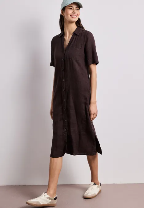 Vrouw draagt een losvallende donkerbruine midi-hemdjurk met korte mouwen, zijsplitsen, beige sneakers en pet.