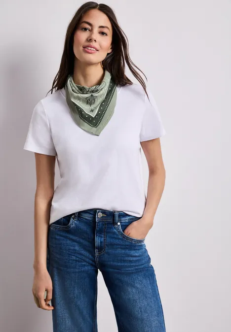 Frau mit weißem T-Shirt, blauer Jeans und grünem Halstuch, eine Hand in der Tasche.