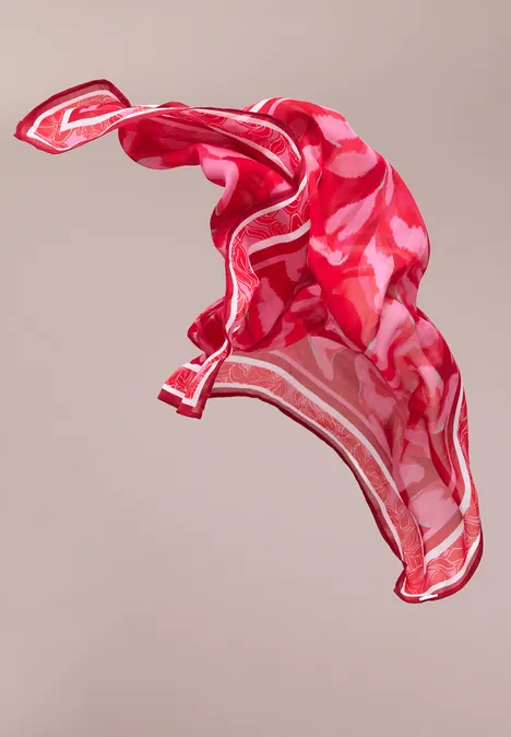Ein rotes, gemustertes Seidentuch schwebt leicht in der Luft vor neutralem Hintergrund.