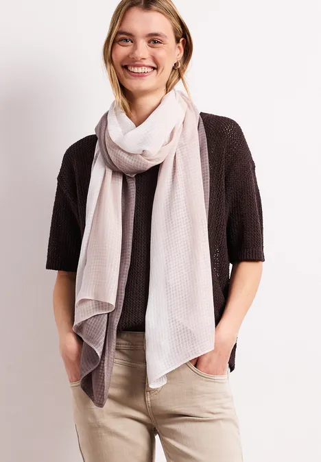 Frau mit hellem Schal, schwarzem Cardigan und beiger Hose, Hände in den Taschen.