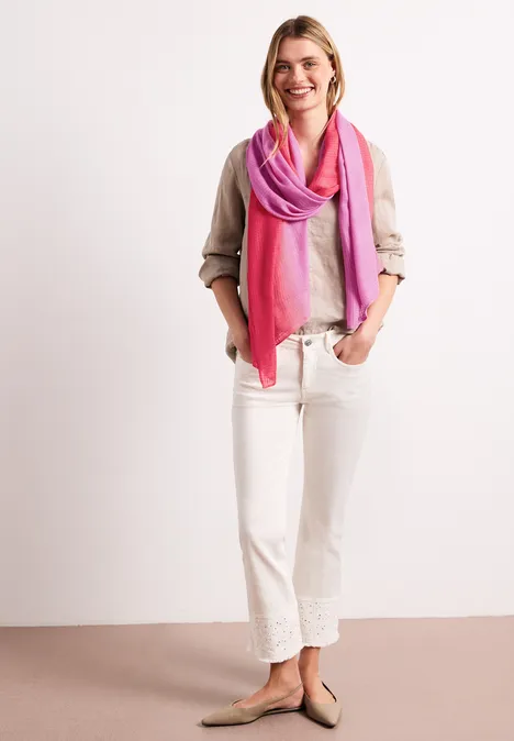Person in beiger Bluse, weißer weiter Hose und pink-lila Schal vor neutralem Hintergrund.