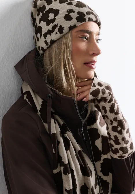 Femme de profil portant bonnet, écharpe et gants à motif léopard devant un mur clair.