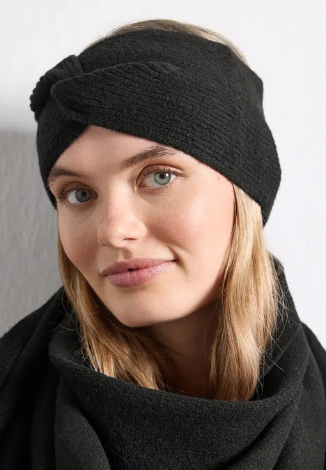 Frau mit hellem Haar trägt schwarzen Strick-Stirnband-Turban und passenden Schal vor weißer Wand.
