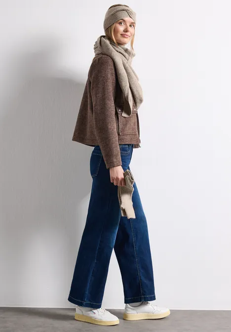 Frau in breiter Jeans, braunem Pullover, Schal und Mütze posiert seitlich vor weißem Hintergrund.