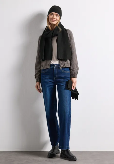 Modische Frau in kaltem Wetter-Outfit: Mütze, Schal, Jacke, Jeans und Stiefel, hält Handschuhe.