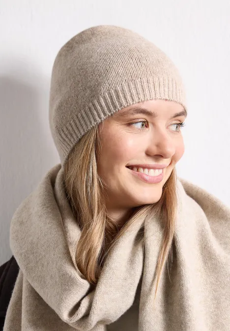 Eine lächelnde Frau trägt eine beige Mütze und einen passenden Schal vor weißem Hintergrund.