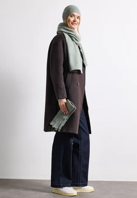 Frau mit Wintermantel, Schal und Mütze, weiten Jeans und Sneakern, lächelnd und hält Clutch
