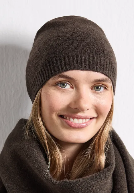 Feinstrick Beanie in Unifarbe auburn brown mel. Junge Frau lächelt in brauner Strickmütze und passendem Schal vor weißem Hintergrund.