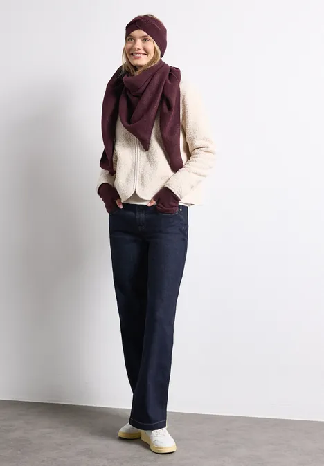 Frau in warmer Herbstmode: helle Strickjacke, lila Mütze, Schal, Jeans und Sneaker. Sie lächelt an der Wand.