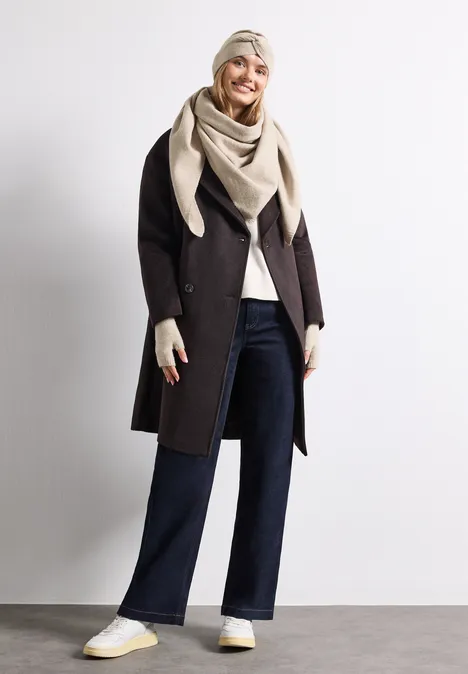 Femme en manteau sombre, jean large, étole crème, gants et baskets blanches sur fond blanc.