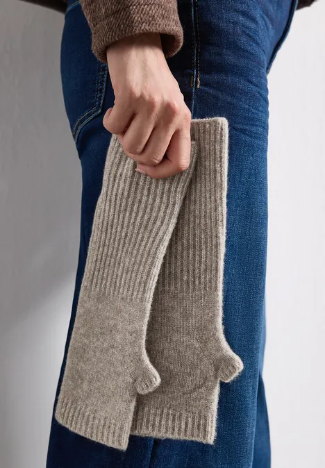 Eine Person in Jeans hält ein Paar graue, gestrickte Stulpen mit Daumenöffnung.