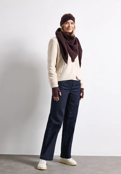 Fijn gebreide armwarmers in effen kleur rustic rouge mel. Vrouw in stijlvolle winteroutfit: lichte trui, bordeaux muts en sjaal, handschoenen, donkerblauwe jeans en witte sneakers.