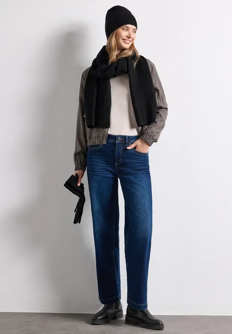 Lange, fijngebreide sjaal in effen kleur Black Vrouw in blauwe jeans, licht shirt, zwarte sjaal en muts, met klein tasje voor een grijze muur