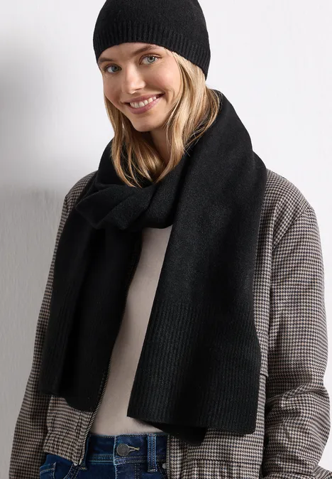 Lange, fijngebreide sjaal in effen kleur Black Jonge blonde vrouw lacht, draagt zwarte muts, zwarte sjaal en geruite jas voor een witte achtergrond.