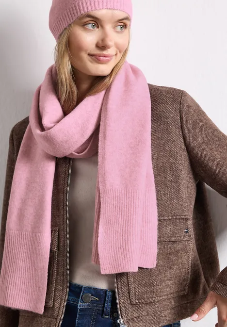 Frau mit rosa Mütze, rosa Schal und brauner Jacke vor hellem Hintergrund.