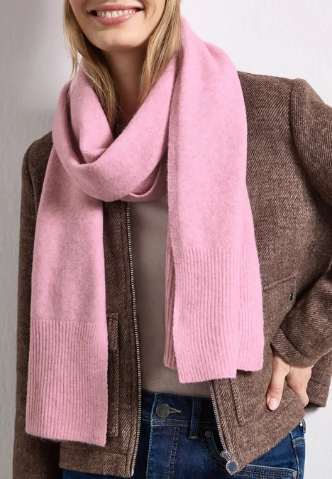 Eine lächelnde Frau mit rosafarbenem Schal, brauner Jacke und Jeans.