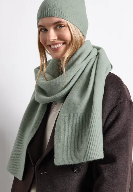 Lange, fijngebreide sjaal in effen kleur eucalyptus green mel. Een glimlachende vrouw draagt een groen gebreide muts en bijpassende sjaal bij een donkere jas.