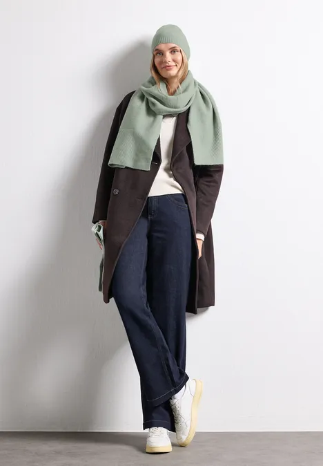 Lange, fijngebreide sjaal in effen kleur eucalyptus green mel. Vrouw met lichtgroene sjaal en muts, donkere jas, wijde jeans en witte sneakers tegen witte achtergrond.