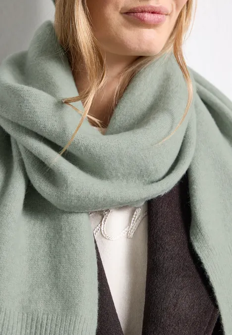 Lange, fijngebreide sjaal in effen kleur eucalyptus green mel. Een vrouw draagt een zachte mintgroene sjaal en een donkere jas over een lichte trui.