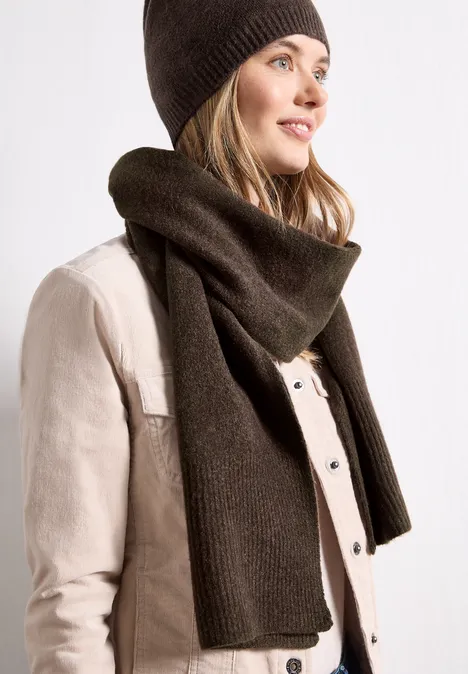 Frau mit brauner Strickmütze und Schal, helle Jacke, lächelnd seitlich blickend.