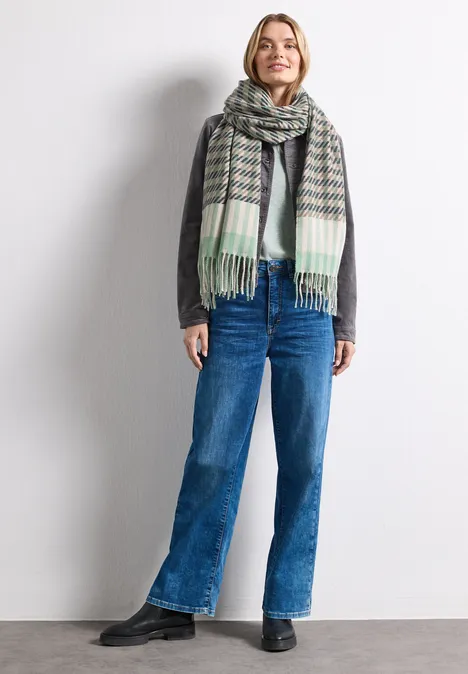 Frau trägt grauen Pullover, große Karoschal, blaue gerade Jeans und lächelt.