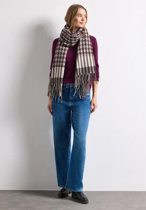 Frau vor weißer Wand mit lila Pullover, kariertem Schal und blauen Jeans, lässig stehend.