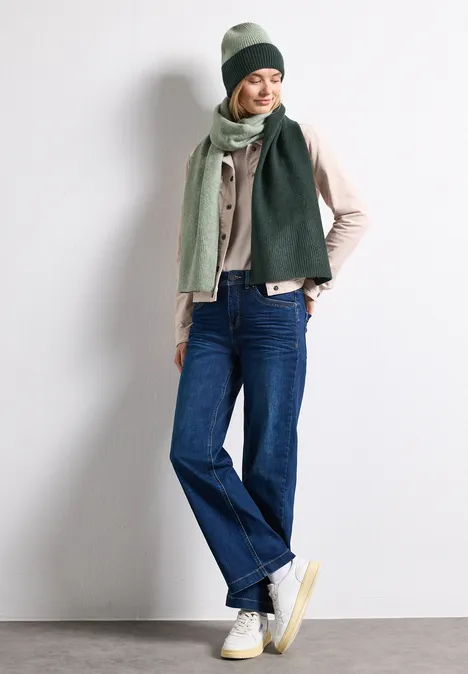 Femme en jean, veste beige, écharpe et bonnet verts, baskets blanches, adossée décontractée.