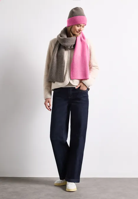 Une femme porte un jean large, un chemisier clair, une écharpe grise et rose et un bonnet assorti devant un fond blanc.