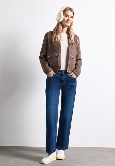 Teddy-Ohrwärmer mit Veloursdetails barley beige Eine lächelnde Frau trägt eine braune Jacke, weißen Pullover, blaue Jeans und weiße Sneaker vor weißem Hintergrund.