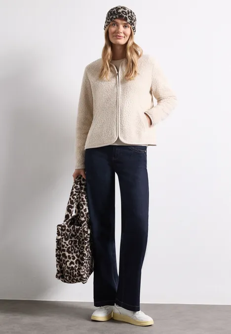 Teddy-Stirnband mit Leo-Muster auburn brown Frau mit beiger Teddyjacke, schwarzer Mütze, dunklen Jeans, weißen Sneakern und Leopardenmuster-Tasche vor weißer Wand.