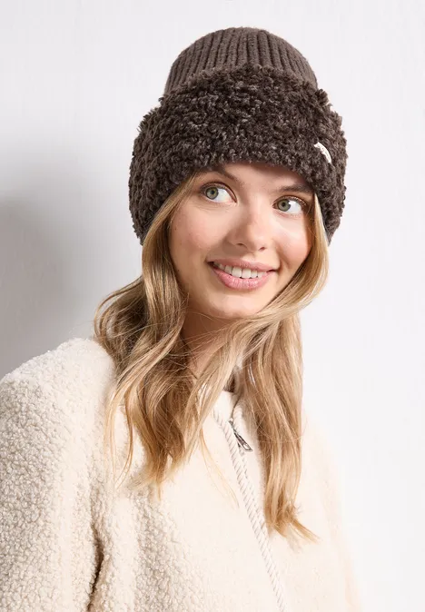 Jonge vrouw lacht, draagt een beige teddyjas en een bruine, pluche gebreide muts.