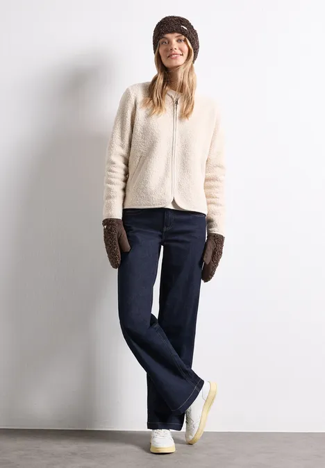 Een jonge vrouw glimlacht in een lichtkleurige gebreide trui, donkere jeans, bruine wanten en een muts.