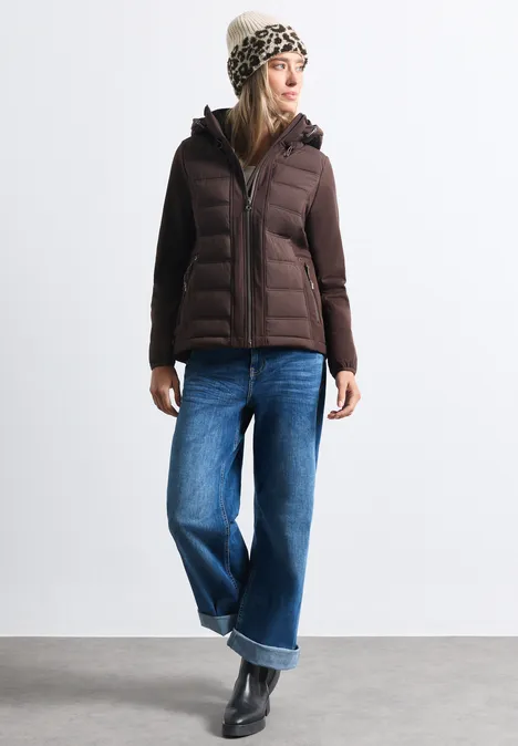 Femme portant une doudoune marron, jean large, bottes noires et bonnet à motif sur fond blanc.