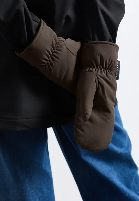 Personne porte une veste noire, un jean bleu et des moufles brunes, gros plan sur fond neutre.