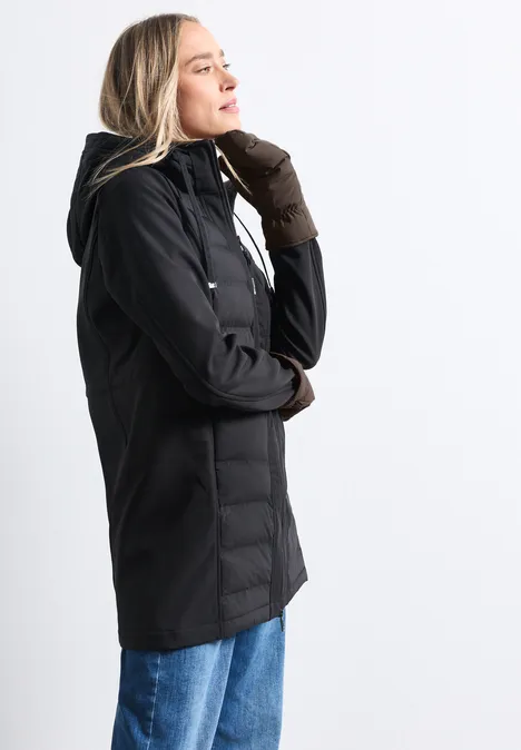 Femme de profil portant une veste noire à capuche, gants marron et jean bleu sur fond clair.