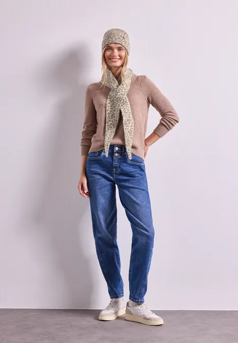Frau in Beanie, Schal, beige Strickjacke, blauen Jeans und Sneakern steht lächelnd vor heller Wand.
