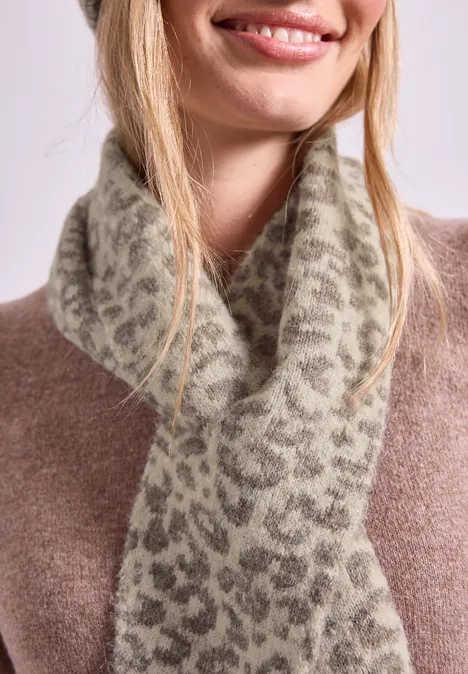 Eine lächelnde Frau trägt einen flauschigen Schal mit Leo-Muster über einem braunen Pullover.