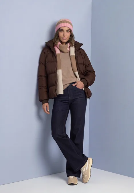 Frau in brauner Daunenjacke, rosa Mütze und Schal, beiges Strickshirt, dunkle Jeans und weiße Sneaker.