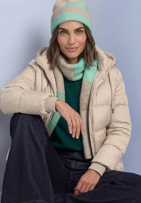 Vrouw in beige donzen jas met groen-beige muts en sjaal, groene trui en donkere broek, glimlachend.