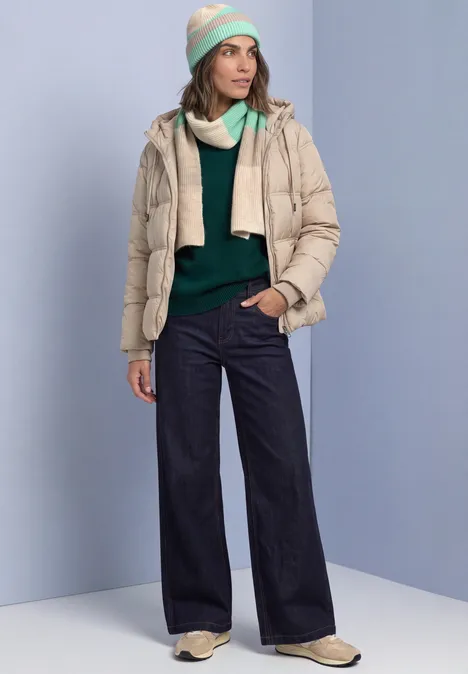 Vrouw in lichtblauwe studio met beige donsjas, groene trui, wijde spijkerbroek, beige laarzen en pastelmuts