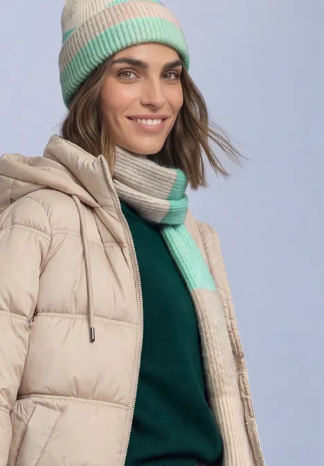 Een vrouw lacht en draagt een beige donzen jas, groene trui, beige-groene sjaal en muts.
