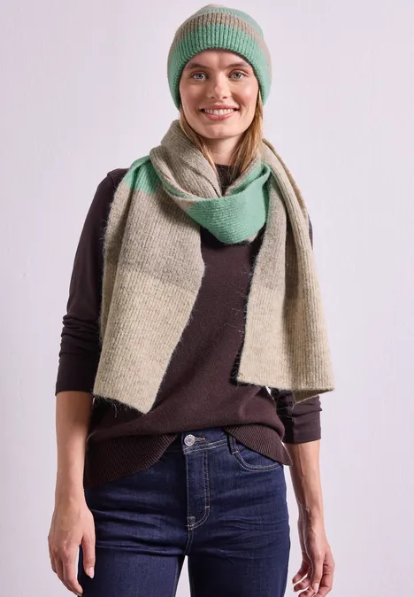 Frau mit grün-grauem Beanie und Schal lächelnd vor hellem Hintergrund.